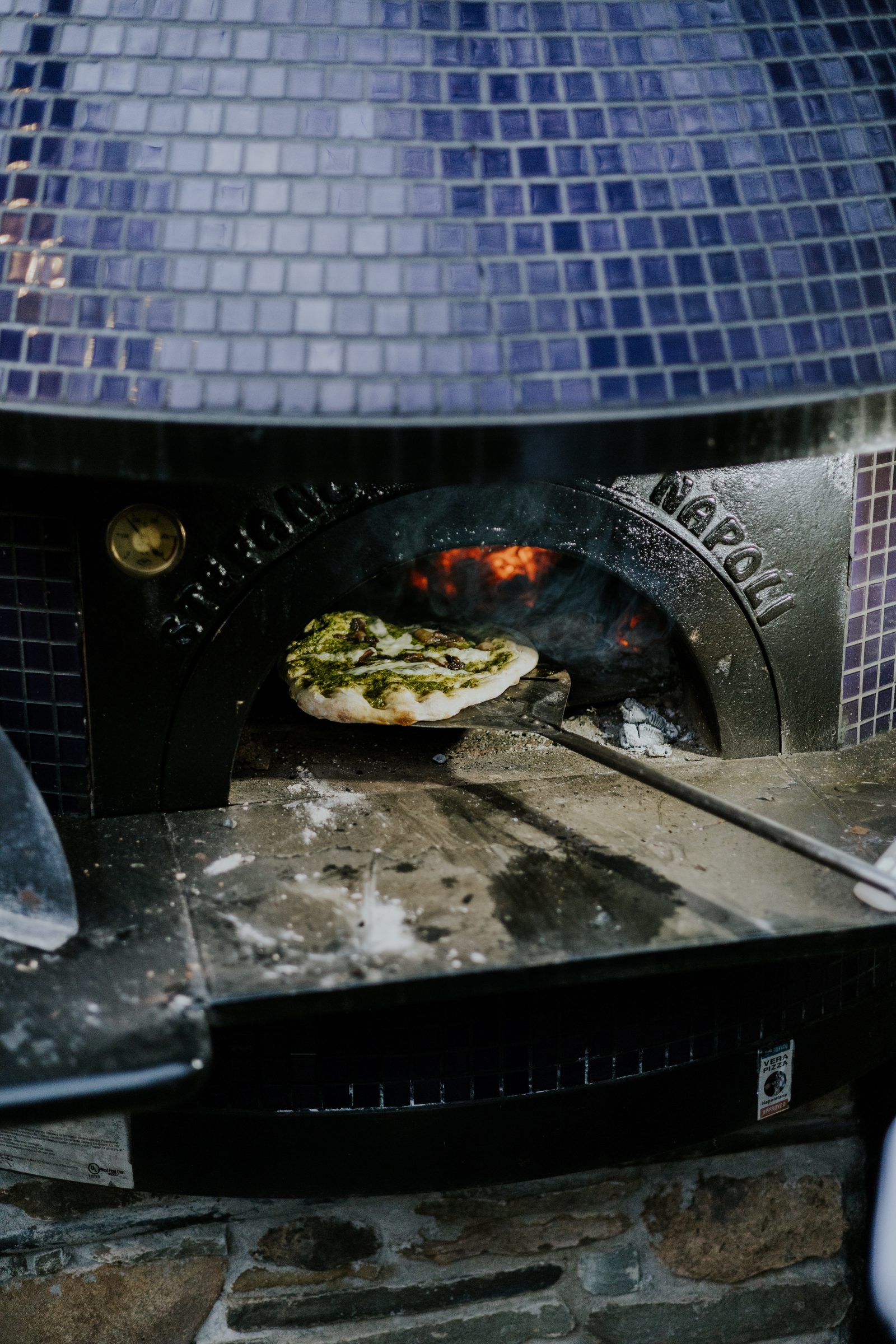 A pesto pizza coming out of the wood-fired oven